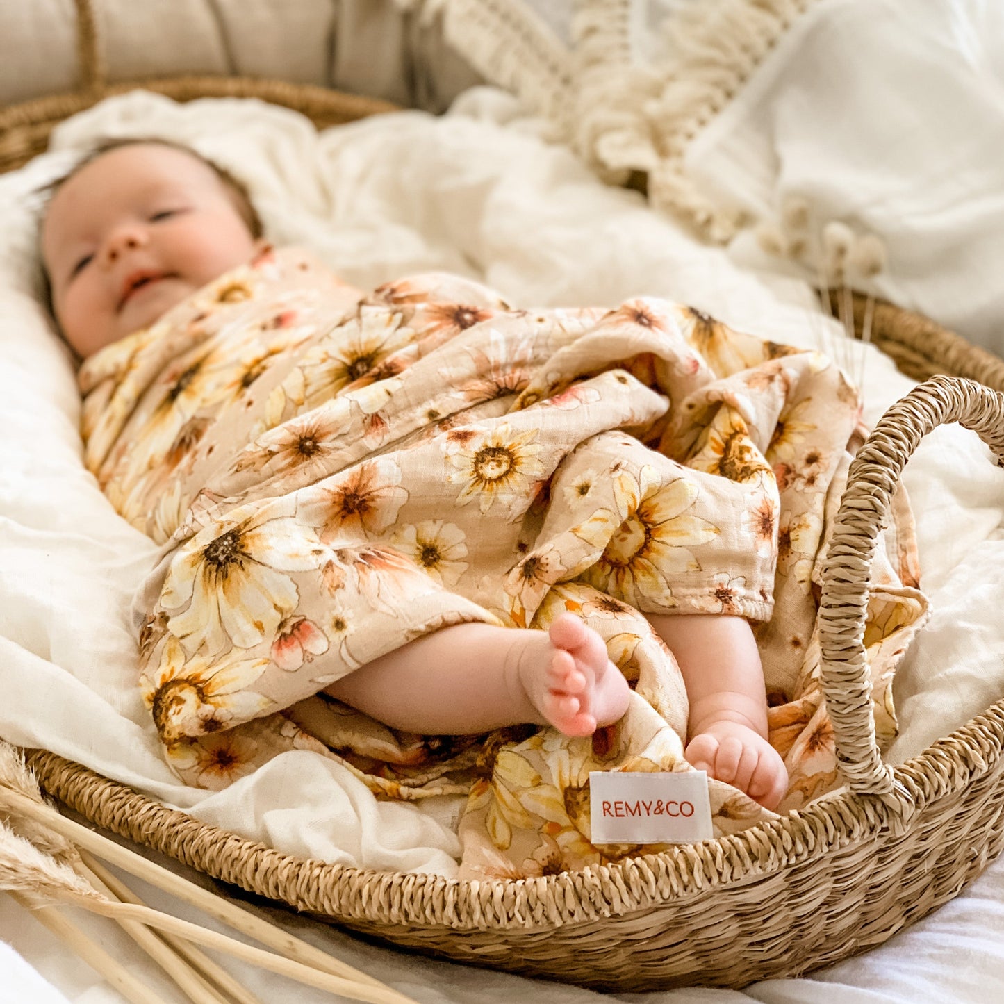 Beautiful 3 month old baby girl swaddled/wrapped in a remy and co floral muslin wrap. Baby is then laying in a moses basket. Another remy and co muslin lays near by, it is cloud white with tassels around edge. cute baby toes popping out end of swaddle.