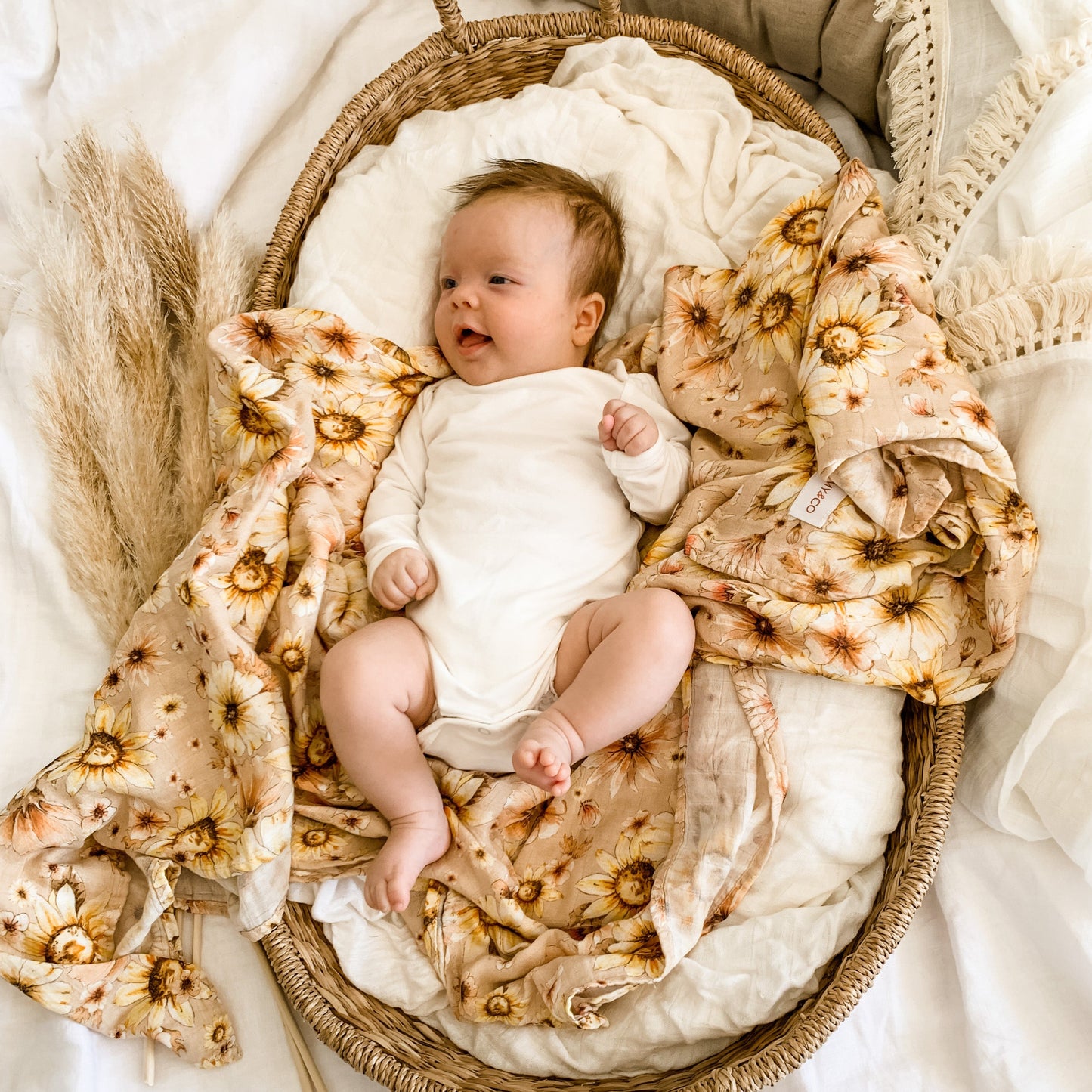 Smiling baby 3months old laying in her moses basket. or bassinet. Beautiful Remy and Co floral muslin wrap draped over baby bed that baby is lying on top of.